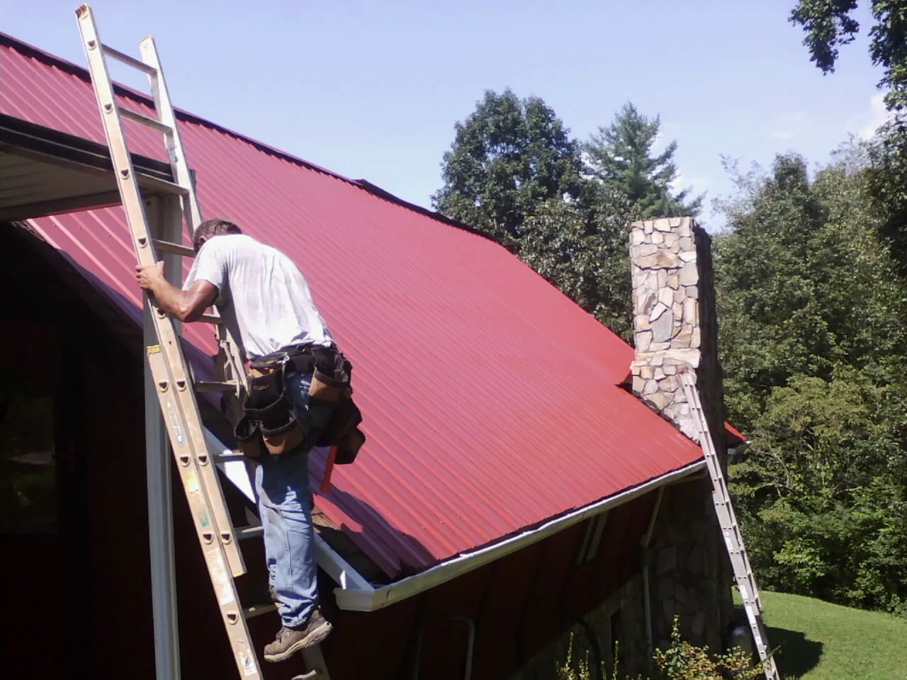 Roofer on a ladder beside a red metal roof and stone chimney — Soffit & Fascia Repair by Roofing Brothers in Tabor